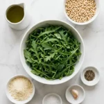 Zutaten für Rucola-Pesto: frischer Rucola, Pinienkerne, Olivenöl, Parmesan und Knoblauch ordentlich aufgereiht
