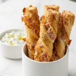 Blätterteig-Schinken-Käse-Stangen serviert mit Tzatziki — krosse Snackstangen perfekt präsentiert neben cremigem Dip